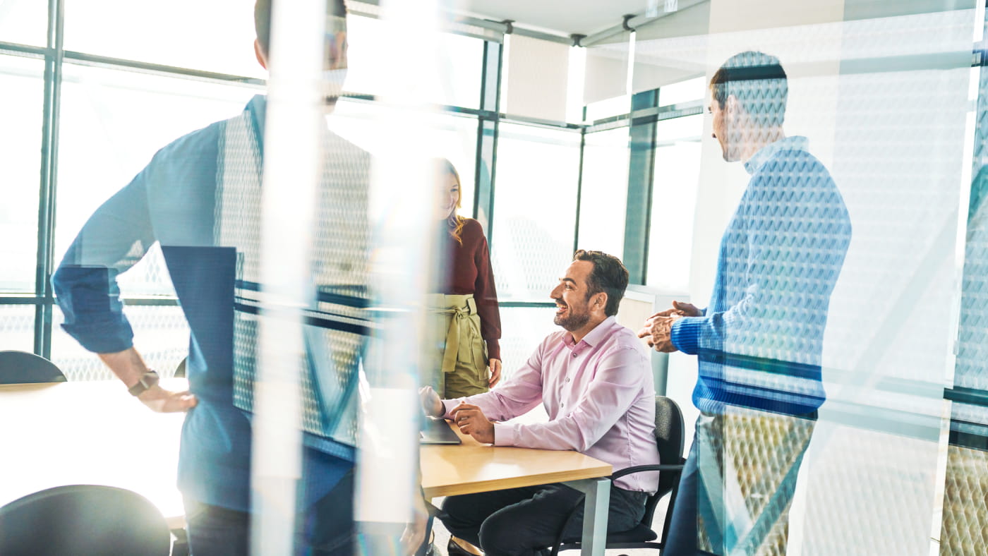 Four people smile and laugh around a table in a bright conference room. Three of them stand at the table while one sits with a laptop on the table in front of them.