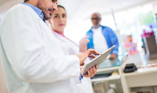 Two pharmacists working on a tablet while a patient is at the counter.