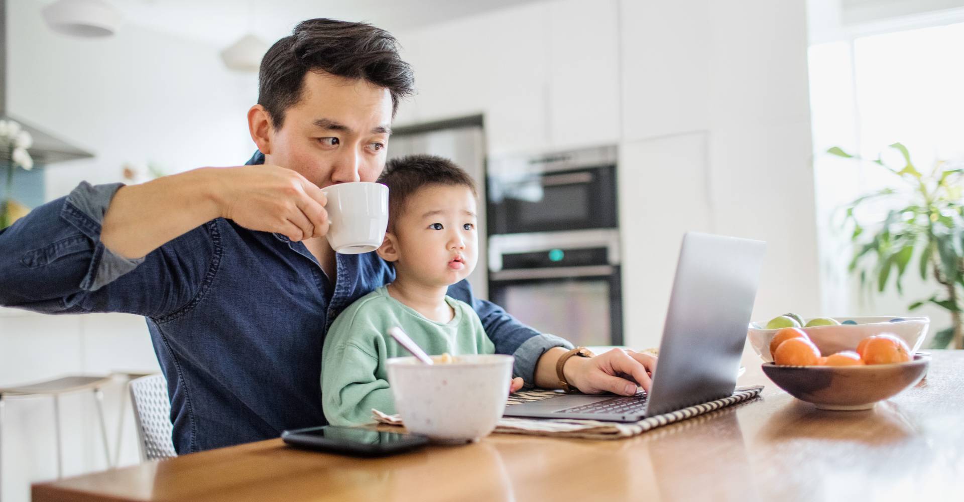 Father multi-tasking with young son at kitchen table