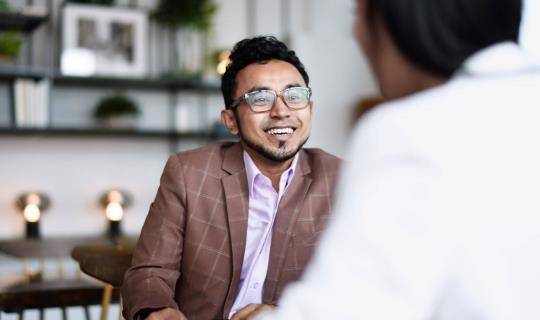 Two people sitting in an office. One of them is facing the camera and smiling